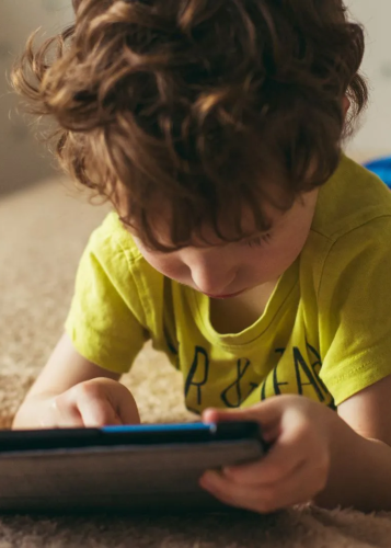 Közeli kép egy göndör hajú kisgyermekről, aki a földön fekve, figyelmesen egy tabletet vagy okostelefont néz. Gyermek, tablet, képernyőidő, digitális média, tanulás, szórakozás, koncentráció, online tartalom, mobil eszköz.